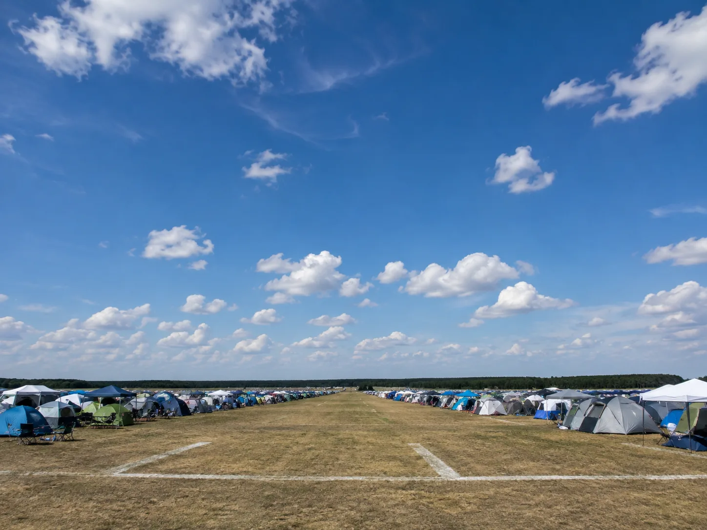 Kampeerterrein aan weerszijden van een brede grasbaan met vervaagde markeringen, tentenrijen tot aan de verre bosrand, weidse blauwe lucht met schapenwolkjes