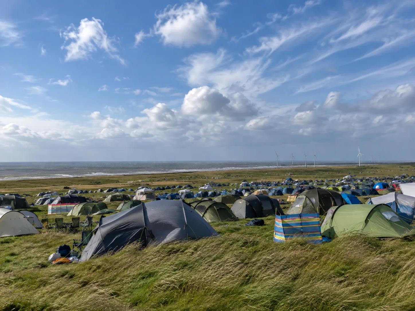 Kampeerterrein op winderig kustgrasland, op de achtergrond de zee en windturbines aan de horizon, gestreepte windschermen tussen de tenten, hoog gras op de voorgrond