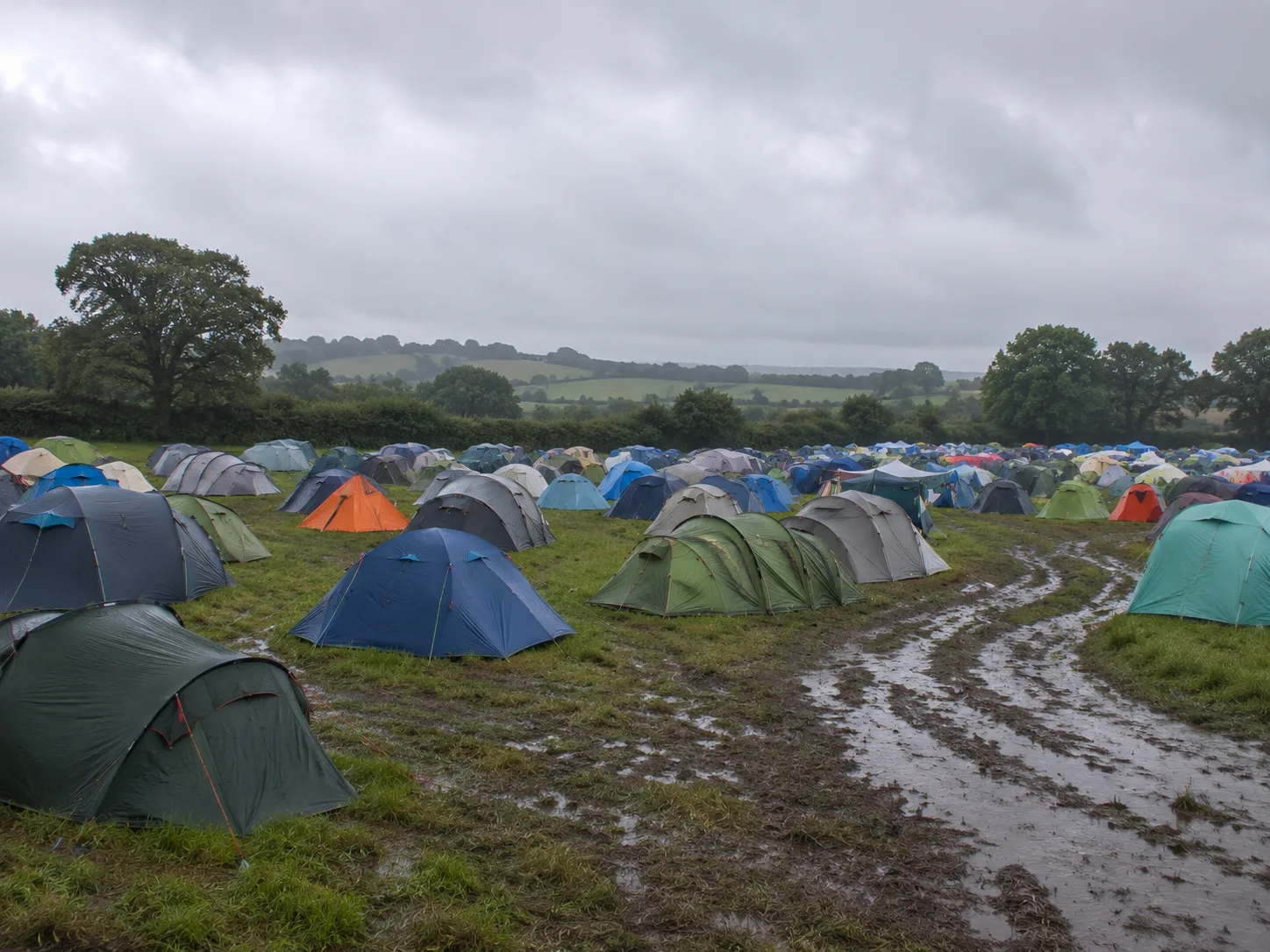 Kampeerterrein op Engels weiland bij regenachtig weer, modderige karrensporen op de voorgrond, heggen en losse eiken aan de veldrand, zachte groene heuvels erachter