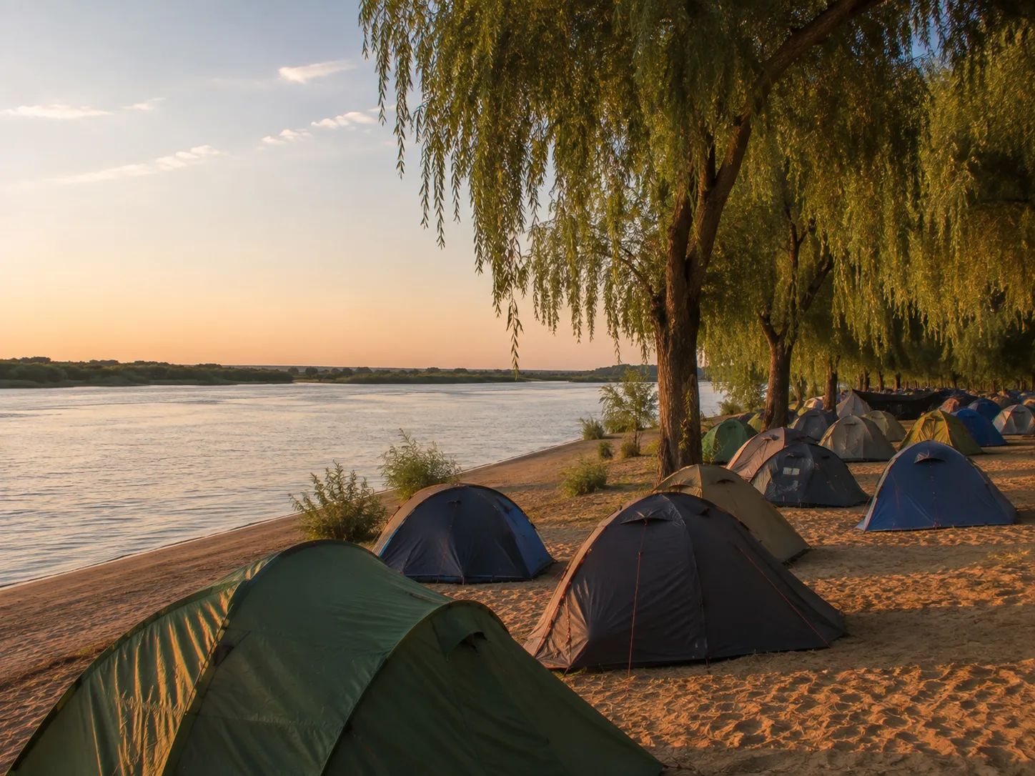 Kampeerterrein op een zandstrand aan de rivier, wilgen langs de oever, warm avondlicht op het zand, een vlakke beboste oever aan de overkant