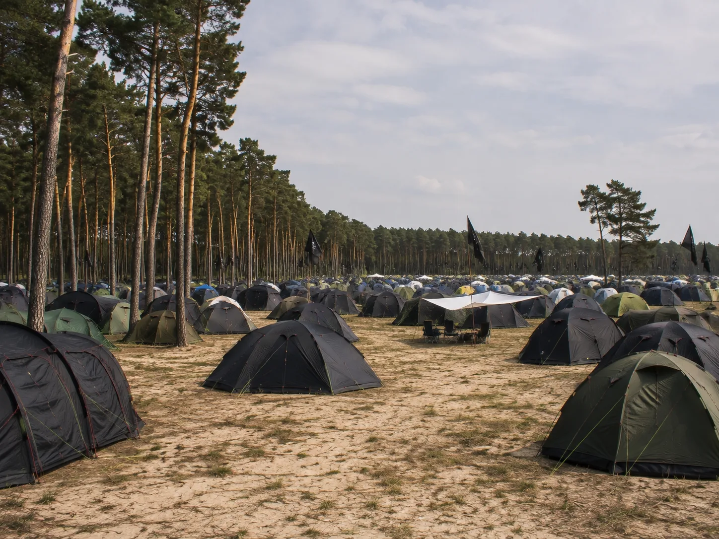 Kampeerterrein op lichte zandgrond naast een hoog dennenbos, donkere tenten en zwarte vlaggen, een wit schaduwdoek met kampeerstoelen eronder