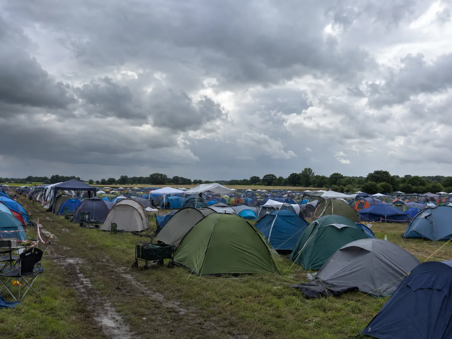 Dicht bezet kampeerterrein op vlak grasland onder donkere regenwolken, een doorweekt karrenspoor op de voorgrond, een bolderwagen tussen de tenten, bomenrij aan de vlakke horizon