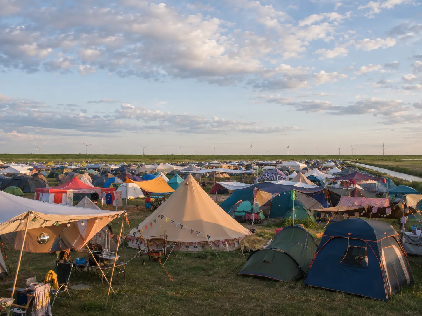 Kleurrijk versierd kampeerterrein in avondlicht, grote tentpaviljoens met lichtslingers en vlaggetjes, daarachter vlak polderland met een vaart en windturbines aan de horizon