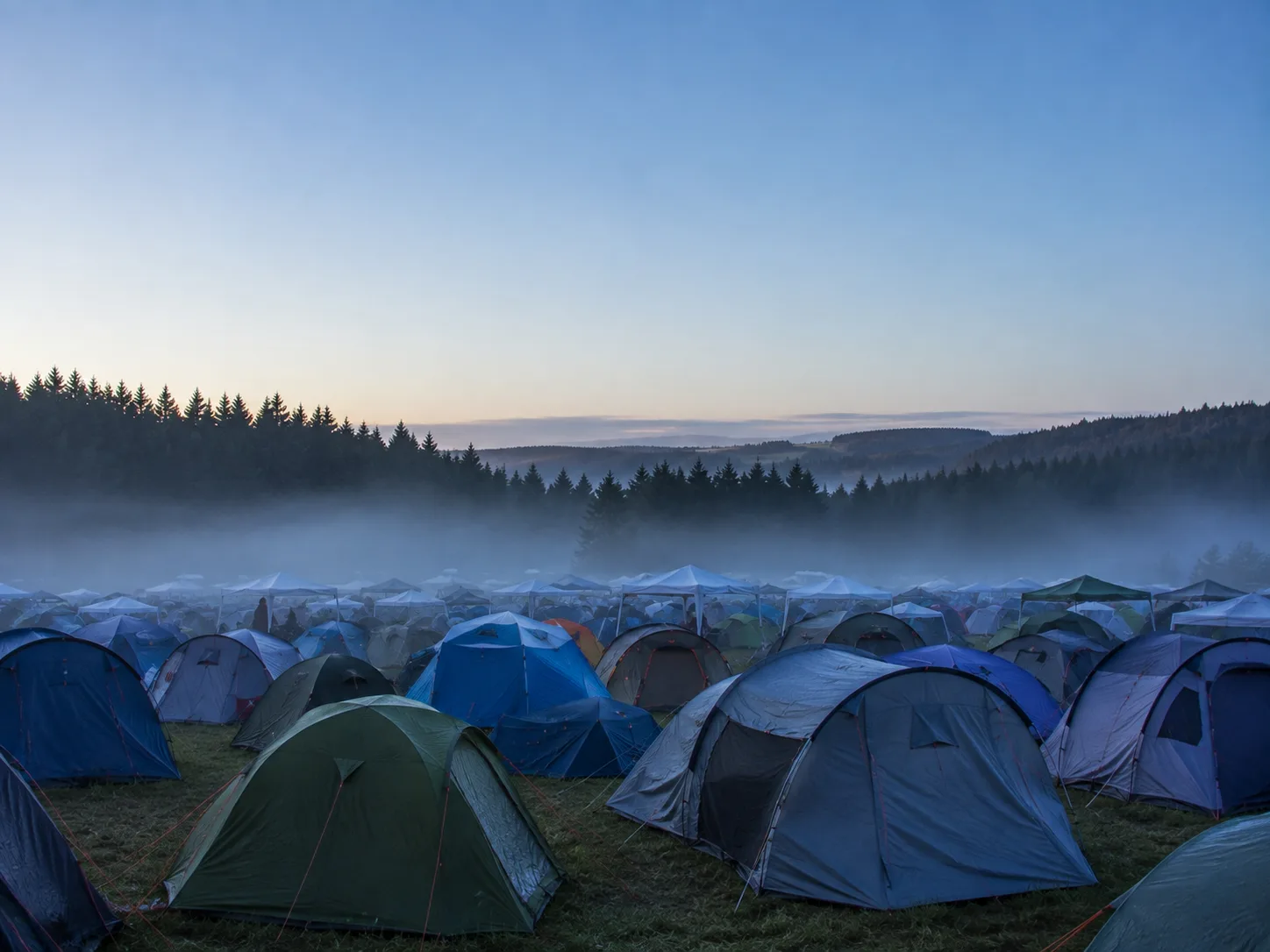 Kampeerterrein op een hoogvlakte bij dageraad, een mistband boven het tentenveld, donkere sparrenbossen en beboste heuvels erachter, koel blauw licht