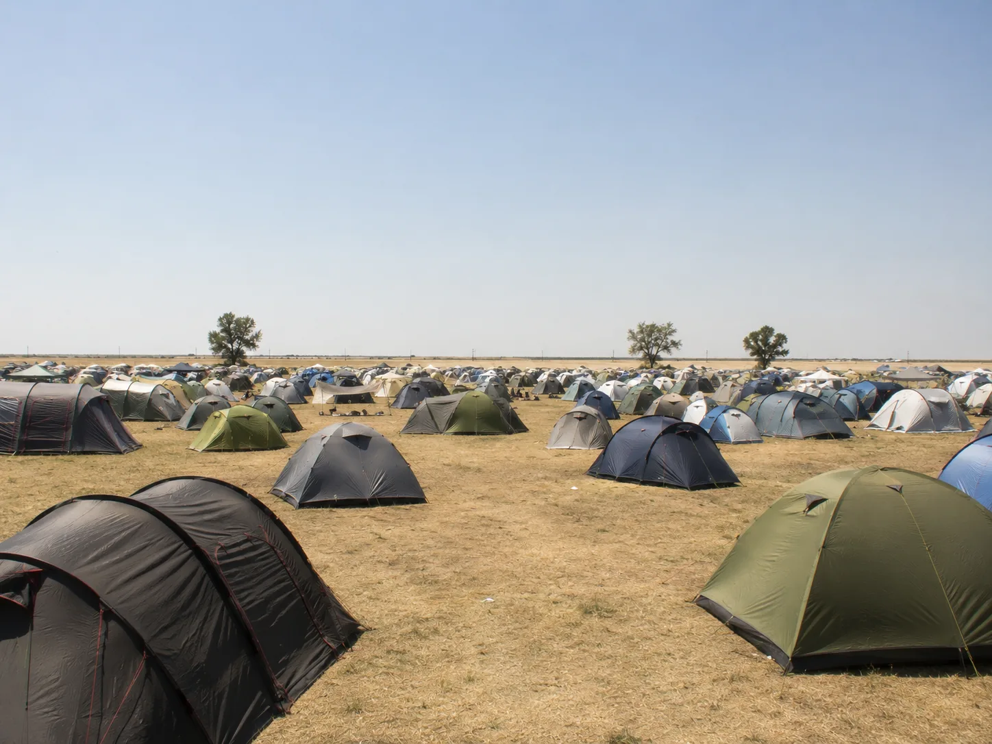 Kampeerterrein op een droge verschroeide vlakte, tenten tot aan de vlakke horizon, slechts enkele losse bomen, bleekblauwe lucht boven trillende hitte