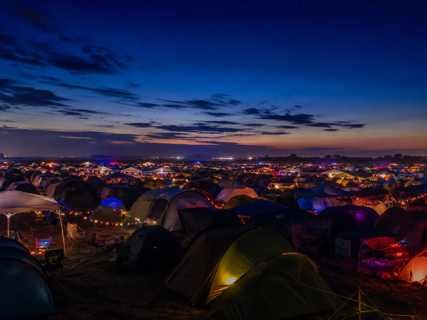 Kampeerterrein in de schemering, honderden tenten tot aan de horizon, warme lichtslingers en gekleurde lampen ertussen, diepblauwe lucht boven een oranje band
