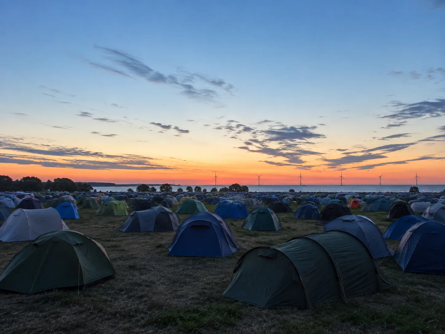 Kampeerterrein op vlak grasland in een lange noordelijke schemering, oranje band aan de horizon boven het water, windturbines in de verte, tenten tot de beeldrand