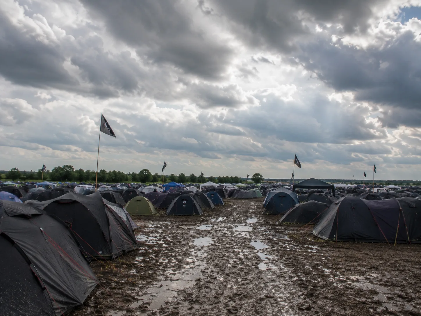Kampeerterrein op vlak marsland onder een weidse bewolkte hemel, donkere tenten langs een omgewoeld modderpad met plassen, losse vlaggen op hoge masten